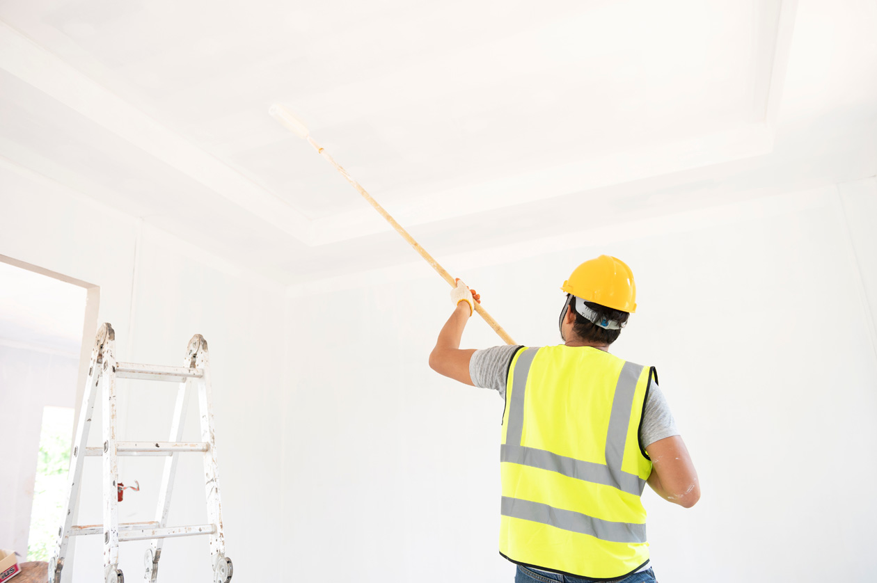 Worker in safety gear painting a ceiling with a roller as part of interior facility maintenance